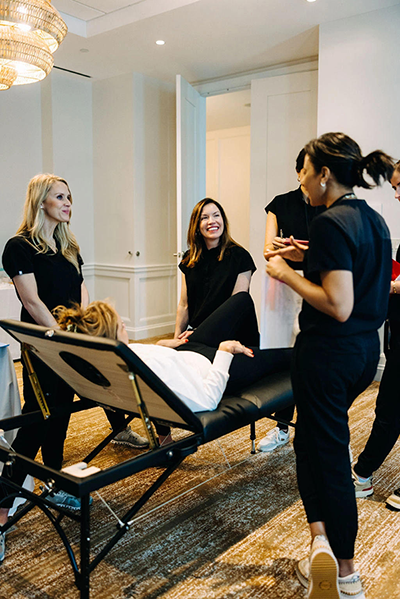 A group of Skin Clique providers gathered and talking around a treatment table during a training session in a hotel room