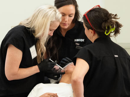 Three Skin Clique providers dressed in black, closely focused as they practise an injection on a live model
