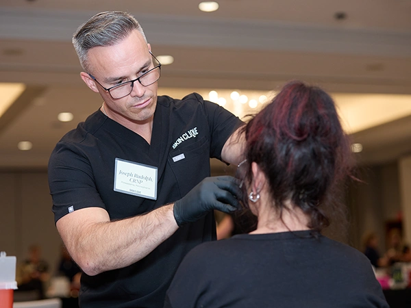 A Skin Clique trainer in black scrubs injecting a seated patient at a training event in a hotel ballroom